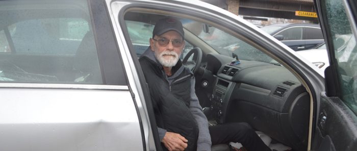 Man sitting in the passenger seat of a car. with the door open, looking at camera.