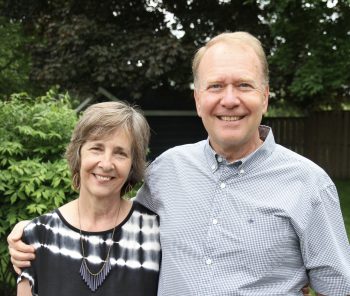 "A smiling older couple stands closely together outdoors in a backyard setting, with green trees and bushes in the background. The woman wears a black and white patterned top and a statement necklace, while the man wears a light blue checkered shirt. They appear happy and relaxed."