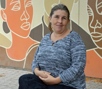 A woman with brown hair tied back sits outdoors in front of a mural with abstract faces. She wears a grey and black striped long-sleeve top and looks calmly at the camera with a gentle smile.