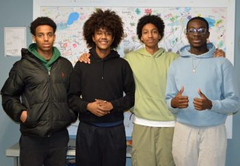 "Four young men stand side by side in front of a colorful, hand-drawn poster. They are smiling and casually dressed in hoodies and sweatpants. One gives a thumbs up, and another has his hand on a friend's shoulder, showing camaraderie."