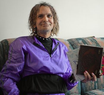 A person with shoulder-length curly hair wearing a shiny purple tracksuit is sitting on a patterned couch, smiling while holding a dark-colored book and some papers. The background includes a second couch with colorful upholstery