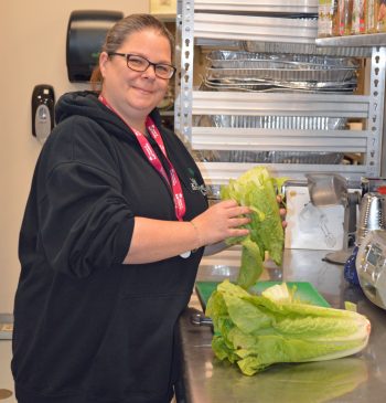 A smiling woman wearing glasses and a black hoodie is standing in a kitchen, preparing romaine lettuce on a stainless steel counter. Behind her are metal shelves with kitchen supplies and a paper towel dispenser on the wall