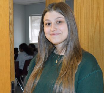 A young woman with long straight hair stands smiling in a doorway, wearing a dark green sweater and a gold necklace. In the background, a group of people is seated in a room with soft lighting from a window